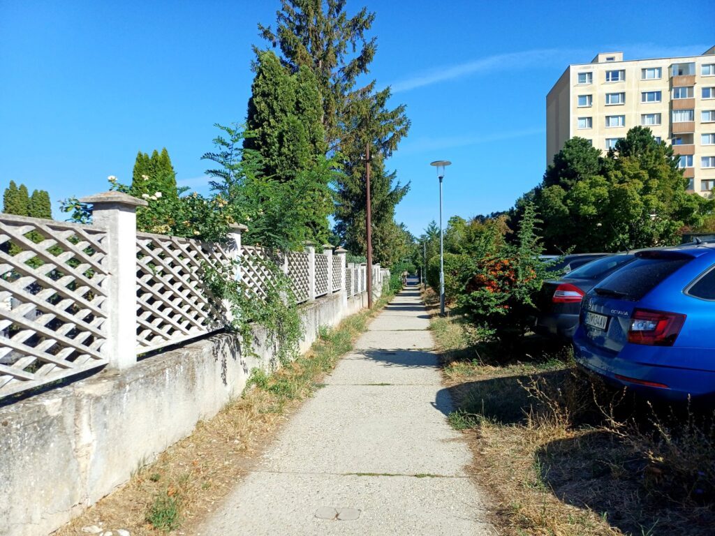 Chodník pri mriežkovom plote cintorína, vedľa zaparkované autá
